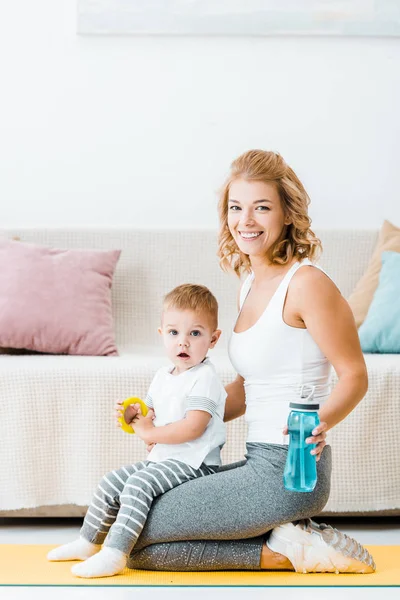 Cute toddler boy and attractive woman sitting on fitness mat and looking at camera at home — Stock Photo