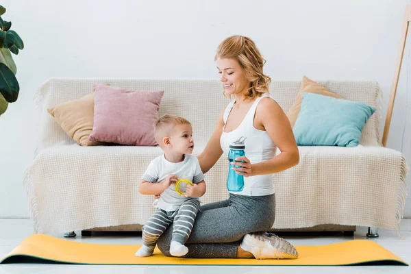 Attrayant femme assise sur tapis de fitness et tenant bouteille de sport et tout-petit garçon avec jouet — Photo de stock
