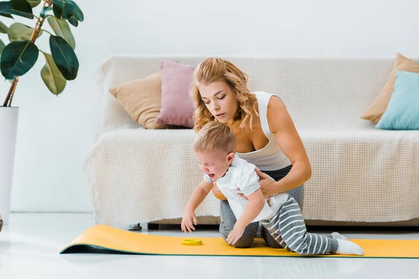 Attractive woman holding crying toddler boy on fitness mat in living room — Stock Photo