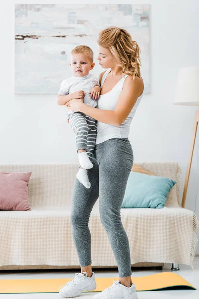 Mère debout près du canapé et tenant tout-petit garçon dans le salon — Photo de stock