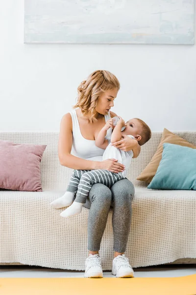 Attractive woman sitting on couch with toddler boy drinking from baby bottle — Stock Photo