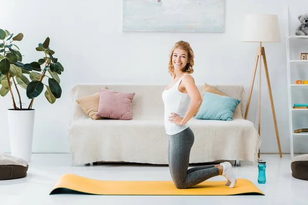 Frau in Sportbekleidung kniet auf gelber Fitnessmatte, blickt in die Kamera und lächelt im Wohnzimmer — Stockfoto