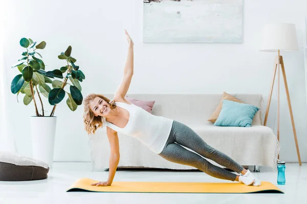 Lächelnde Frau in Sportbekleidung, die auf Fitnessmatte trainiert und im Wohnzimmer in die Kamera schaut — Stockfoto