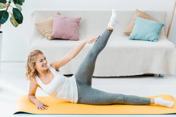 Femme souriante en vêtements de sport étirant la jambe sur tapis de fitness jaune dans le salon — Photo de stock