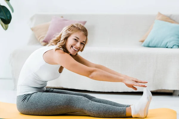 Mujer en ropa deportiva ejercitándose en la alfombra de fitness amarilla, mirando a la cámara y sonriendo en la sala de estar - foto de stock