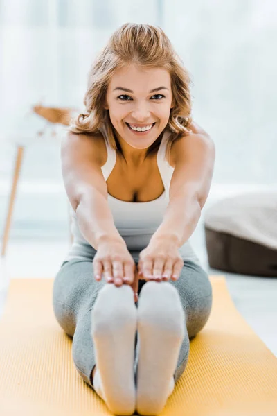 Mujer sonriente haciendo ejercicio de estiramiento en la alfombra de fitness amarillo y mirando a la cámara en casa - foto de stock