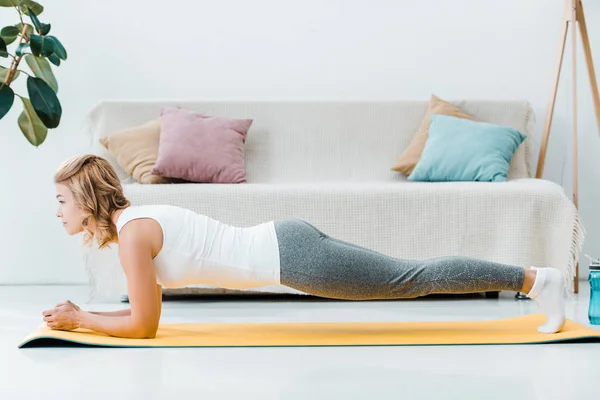 Mujer atractiva en ropa deportiva haciendo tablón en la estera de fitness amarilla en la sala de estar — Stock Photo