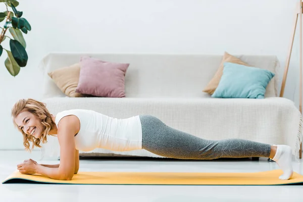 Lächelnde Frau in Sportbekleidung beim Planken auf gelber Fitnessmatte zu Hause — Stockfoto