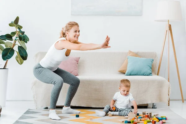 Attractive mother doing squats and cute toddler son sitting on carpet and playing with multicolored cubes at home — Stock Photo