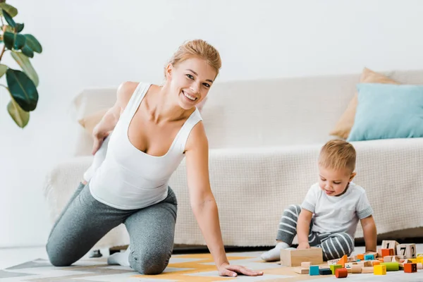 Приваблива жінка робить розтягування вправи на килимі і малюка хлопчика, який грає з різнокольоровими дерев'яними кубиками у вітальні — стокове фото