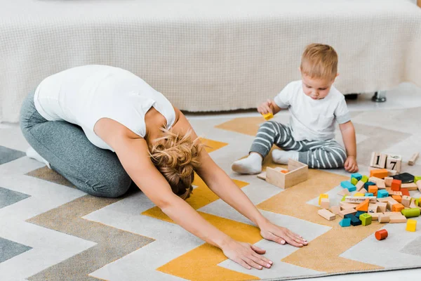 Mulher fazendo exercício de alongamento no tapete e menino brincando com cubos de madeira multicoloridos na sala de estar — Fotografia de Stock