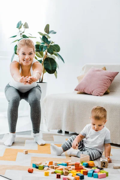 Attraktive Mutter macht Kniebeugen und schaut in die Kamera und niedlichen Kleinkindsohn sitzt auf Teppich und spielt mit bunten Würfeln im Wohnzimmer — Stockfoto
