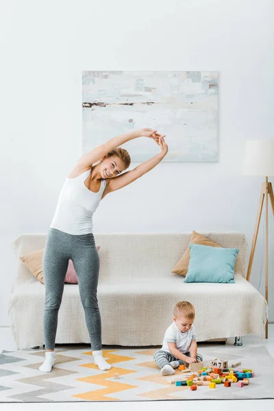 Mujer sonriente estirándose y mirando a la cámara y niño jugando con cubos multicolores en la alfombra en la sala de estar - foto de stock