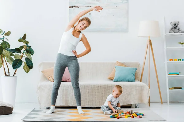 Mujer sonriente haciendo ejercicio y mirando a la cámara y niño jugando con cubos multicolores en la alfombra en la sala de estar - foto de stock