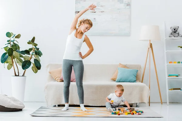 Улыбающаяся женщина делает упражнения и смотрит на милого малыша, играющего с разноцветными кубиками на ковре в гостиной — стоковое фото