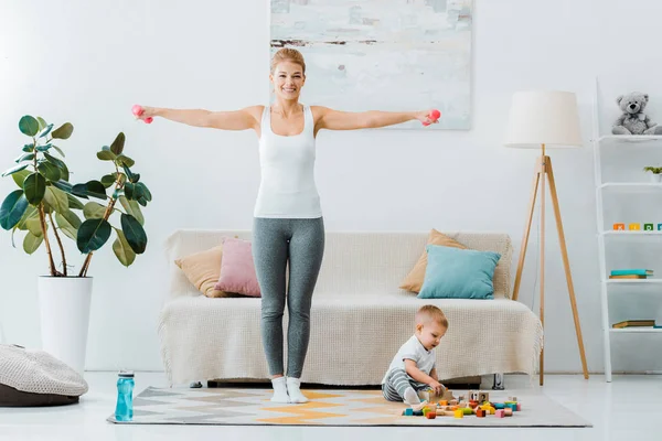 Mujer sonriente haciendo ejercicio con pesas y mirando a la cámara y niño jugando con cubos multicolores en la alfombra en la sala de estar - foto de stock