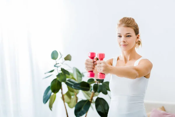 Atractiva mujer en ropa deportiva haciendo ejercicio con mancuernas en casa - foto de stock