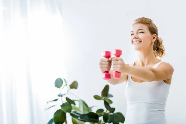 Femme attrayante dans les vêtements de sport souriant et faire de l'exercice avec des haltères à la maison — Photo de stock