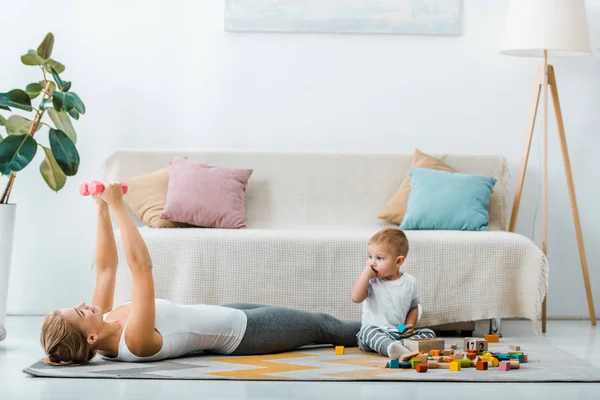 Mujer acostada en la alfombra y haciendo ejercicio con pesas y lindo niño sentado cerca de cubos multicolores de madera en la sala de estar - foto de stock