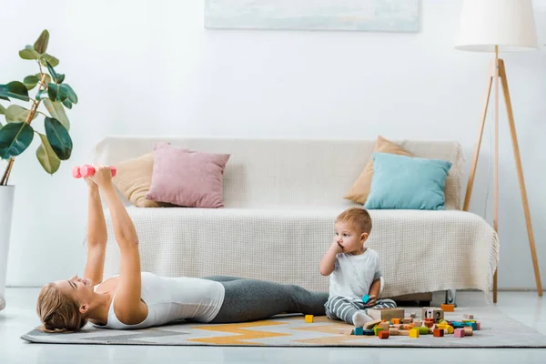 Femme couchée sur le tapis et l'exercice avec des haltères et mignon tout-petit garçon assis près de cubes multicolores en bois dans le salon — Photo de stock