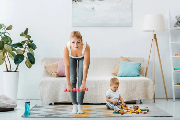 Attraktive Frau macht Übungen mit Kurzhanteln und schaut in die Kamera mit niedlichem Kleinkind, das im Wohnzimmer mit bunten Würfeln spielt — Stockfoto