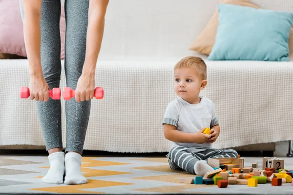 Abgeschnittene Ansicht einer Frau beim Fitnesstraining mit Kurzhanteln und einem niedlichen Kleinkind, das auf einem Teppich neben bunten Würfeln sitzt — Stockfoto