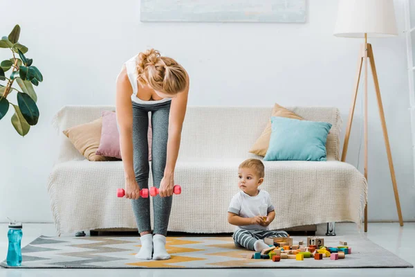 Femme attrayante faisant de l'exercice avec des haltères et en regardant mignon tout-petit garçon assis sur le tapis près de cubes multicolores dans le salon — Photo de stock