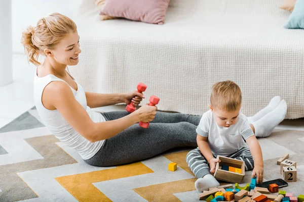 Attraente donna esercizio con manubri e guardando carino bambino ragazzo in possesso di scatola di legno con cubi multicolori vicino al divano — Foto stock