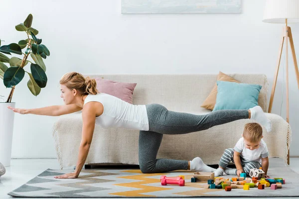 Mulher exercitando perto de sofá e criança menino brincando com cubos de madeira multicoloridos no tapete na sala de estar — Fotografia de Stock
