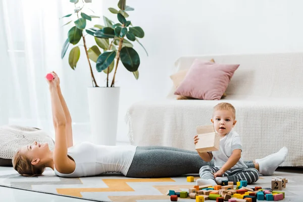 Attractive woman lying on carpet and doing exercise with dumbbells and cute toddler boy holding wooden box in living room — Stock Photo