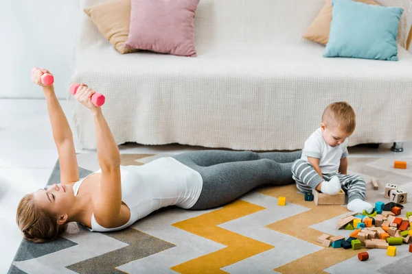 Приваблива жінка робить вправи з гантелями і милий хлопчик-малюк грає з різнокольоровими дерев'яними кубиками на килимі у вітальні — стокове фото