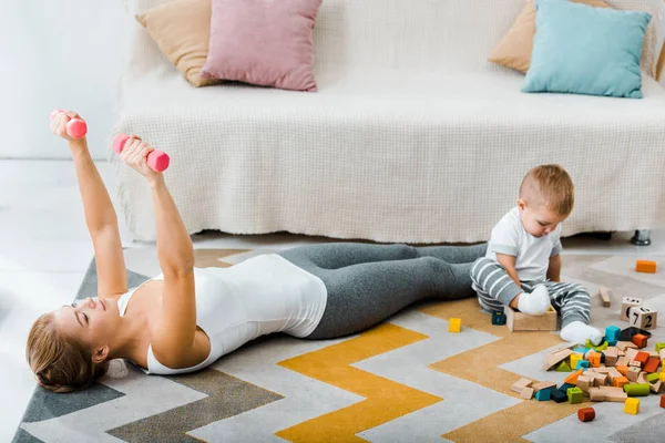 Attractive woman doing exercise with dumbbells and cute toddler boy playing with multicolored wooden cubes on carpet in living room — Stock Photo