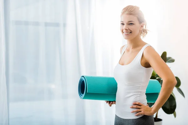 Mulher atraente sorrindo e segurando tapete de fitness na sala de estar — Fotografia de Stock
