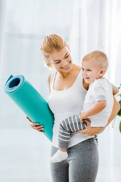 Attractive woman in sportswear holding fitness mat and smiling toddler boy — Stock Photo