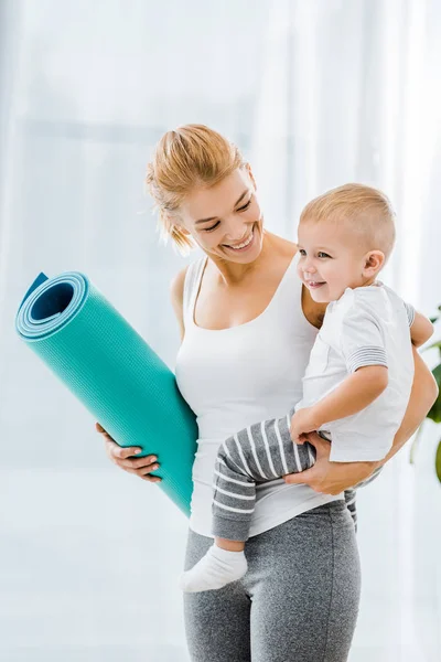 Atractiva mujer en ropa deportiva celebración de la alfombra de fitness y sonriente niño - foto de stock