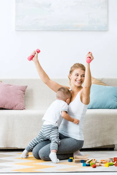 Smiling woman holding dumbbells in raising hands with toddler boy embracing mother in living room — Stock Photo