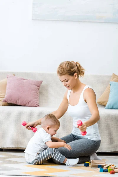 Mulher atraente segurando halteres com menino criança sentado no tapete perto do sofá na sala de estar — Fotografia de Stock