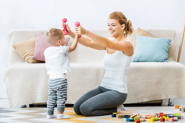 Smiling woman holding dumbbells in raising hands with standing toddler boy on carpet in living room — Stock Photo