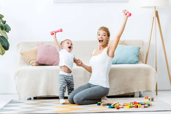 Madre riendo y niño pequeño sosteniendo pesas en levantar las manos y mirando la cámara en la alfombra en la sala de estar - foto de stock