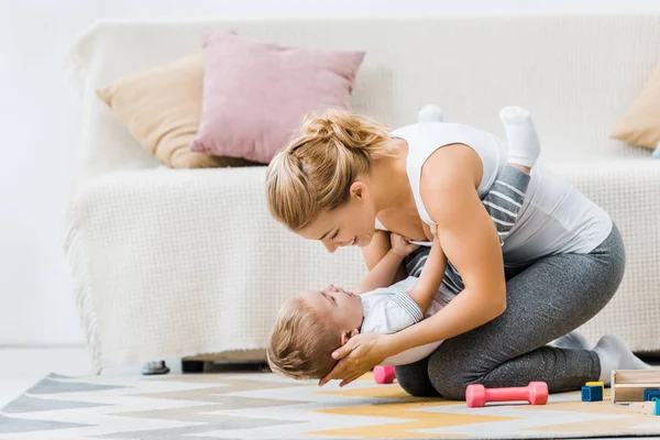 Lächelnde Frau spielt mit entzückendem Kleinkind auf Teppich im Wohnzimmer — Stockfoto