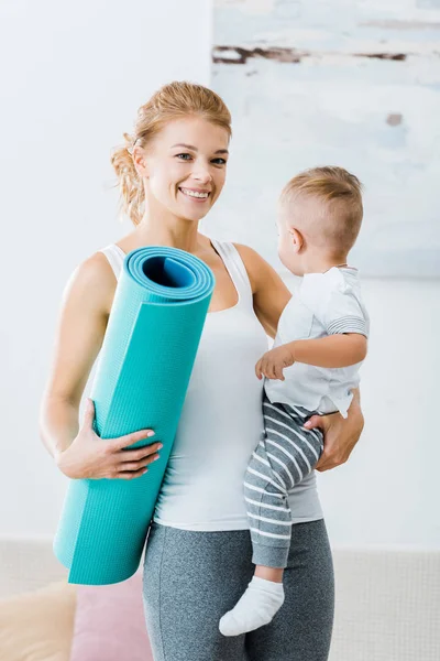 Sonriente mujer sosteniendo la alfombra de fitness y niño pequeño y mirando a la cámara en casa - foto de stock