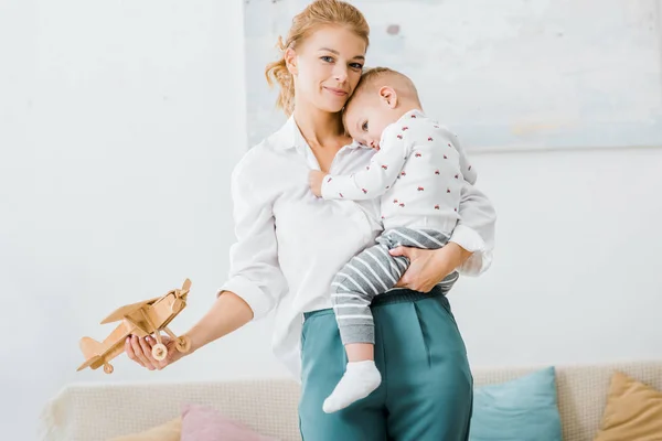 Attrayant femme regardant caméra et tenant modèle d'avion en bois et tout-petit garçon dans le salon — Photo de stock