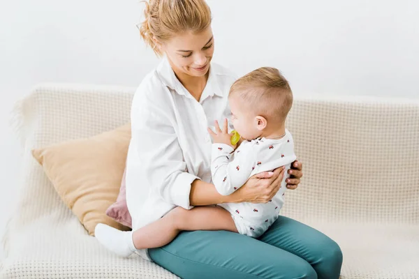 Frau sitzt auf Sofa, hält Kleinkind-Sohn und lächelt im Wohnzimmer — Stockfoto