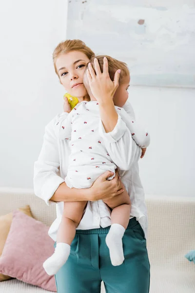 Madre che tiene in braccio il figlio del bambino e guarda la fotocamera in soggiorno — Foto stock