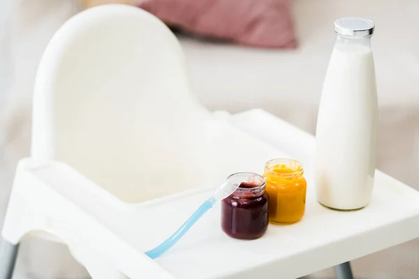 Baby food jars with spoon and milk bottle on white highchair in living room — Stock Photo
