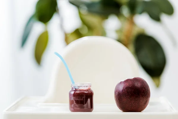 Vista de cerca de la manzana roja y el frasco de alimentos para bebés con cuchara en la trona - foto de stock