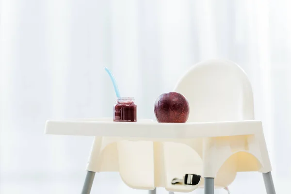 Maçã vermelha e comida de bebê em frasco com colher em cadeira alta branca — Fotografia de Stock