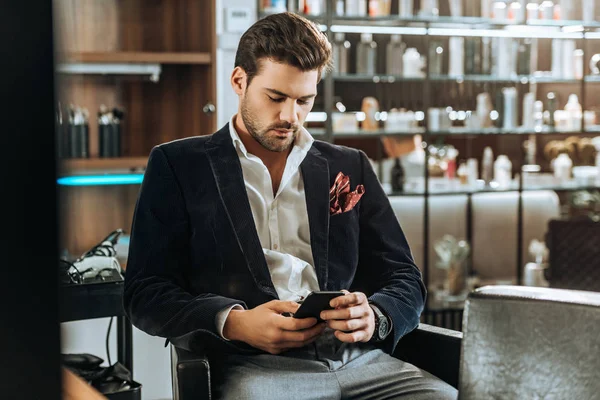 Joven guapo elegante usando el teléfono inteligente mientras está sentado en el salón de belleza - foto de stock