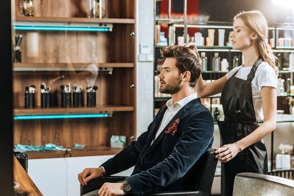 Vista lateral de peluquero joven haciendo peinado al cliente masculino guapo en salón de belleza - foto de stock