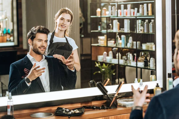 Joven peluquero sonriente y cliente masculino mirando el espejo en el salón de belleza - foto de stock
